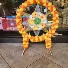 A beautiful circular Ganapati backdrop frame decorated with artificial marigold garlands in yellow and orange. The center features a star-shaped design with gerbera daisies, creating a traditional and auspicious look.