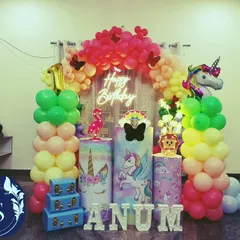 A complete unicorn party stage with a rainbow balloon arch and themed plinths. The setup is brightened by a neon sign and includes marquee letters spelling out the birthday girl's name, creating a perfect fantasy setting.