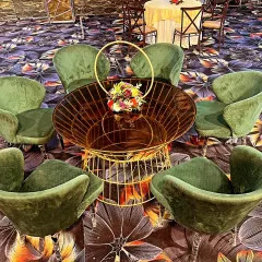 A modern lounge seating cluster with plush green velvet chairs. The centerpiece is a unique gold wire-frame table with a small floral arrangement, perfect for cocktail hours.