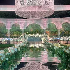 A stunning reflective aisle mirrors the grand, illuminated floral chandelier above, creating a magical pathway for the couple. The aisle is lined with lush white roses and greenery, leading to a stage set against a wall of living plants.