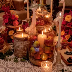 A rich and moody tablescape perfect for a wine and cheese evening. The decor features an abundance of candles, fresh fruits like apples and grapes, and deep red florals, creating a warm, rustic, and opulent still life.