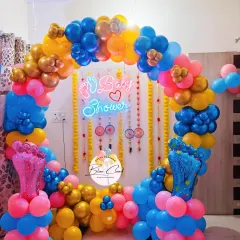 A vibrant baby shower setup with a colorful balloon arch and traditional marigold garlands. The decor features a neon sign and baby feet foil balloons.