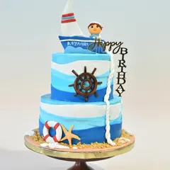 Anchors aweigh for a first birthday. This two-tier sailor-themed cake features blue and white ombre waves, edible sand, and charming nautical details like a ship's wheel, lifebuoy, and a little sailor in his boat.