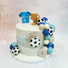 We score a goal with this stylish football champion cake. A personalized jersey, a golden trophy, and a cascade of footballs and spheres in team colors adorn this modern cake, making it the perfect centerpiece for a football fan's birthday.