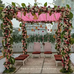 Set against the serene white walls of a temple, this wedding mandap combines tradition with elegance. The four pillars are wrapped in dense floral garlands, while a soft pink fabric canopy and hanging brass bells create a divine atmosphere for the couple.