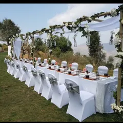 An elegant outdoor dining setup with a long table under a bamboo canopy decorated with white drapes and flowers, perfect for an intimate pre-wedding dinner with a view.