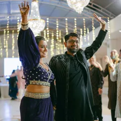 The bride and groom make a grand entrance, waving to their guests at the Sangeet. Their coordinated outfits in shades of blue and black match the event's decor.