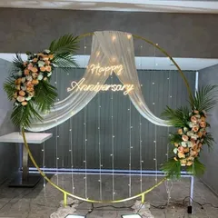 This elegant circular backdrop is perfect for anniversaries or engagements. The golden ring is draped with sheer white fabric and fairy lights, accented with two beautiful floral arrangements and a 'Happy Anniversary' neon sign.