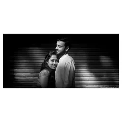 An anniversary portrait using dramatic lighting from window shutters. The black and white treatment emphasizes the texture of the light and the couple's comfortable, loving embrace.