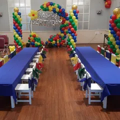 A colorful indoor party setup with long tables for kids and a heart-shaped balloon backdrop in primary colors.