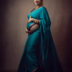 A beautiful studio maternity portrait of a woman in a teal saree, looking at the camera with a gentle smile.