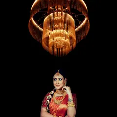 An elegant bridal portrait framed by a magnificent chandelier. The warm, golden light complements her traditional red saree and gold jewelry, creating a look of regal sophistication.