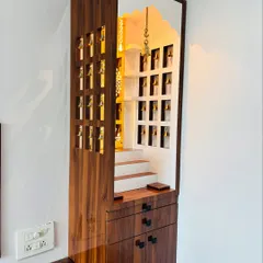A full view of a custom wooden mandir with intricate side panels featuring bell cutouts. The unit includes drawers and cabinets at the base for storing puja items.