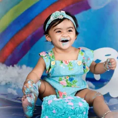 That "I can't believe this is all for me" smile! This little girl is loving every moment of her rainbow-themed cake smash, with blue frosting and a whole lot of happiness.