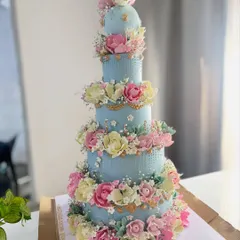A stunning five-tier wedding cake in a soft powder blue. The entire cake is covered in cascading, handcrafted sugar flowers in shades of pink, yellow, and white, creating a romantic and breathtaking effect.