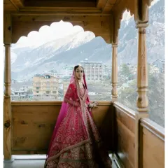 The bride, framed by a traditional wooden balcony, looks out towards the Manali valley. Her vibrant pink lehenga is a stunning splash of color against the earthy tones of the mountains and architecture.