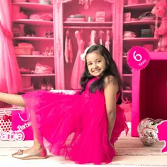 A six-year-old girl smiles brightly in her Barbie-themed shoot, sitting beside a life-sized Barbie box in a room filled with pink.