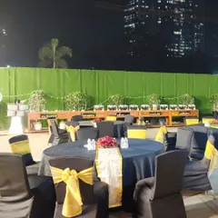 An outdoor reception setup at night. The tables are dressed in black and yellow, with the city skyline visible in the background, creating a chic and urban atmosphere for the celebration.