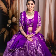 A classic seated bridal portrait. The composition is calm and elegant, allowing the intricate details of the purple lehenga and the gold jewelry to be the main focus.