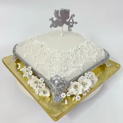 This heavenly angel-themed cake is perfect for a christening or baptism. The square pillow-shaped cake is decorated with intricate white lace piping, silver accents, and a cascade of white sugar flowers, topped with a silver cupid.