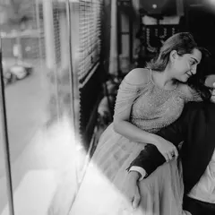 A black and white photo capturing a quiet moment by a large window. The reflection and soft light add an artistic and introspective feel to this urban pre-wedding portrait.