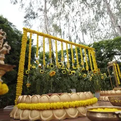 A beautiful Haldi setup designed to be one with nature. The backdrop is made of fresh sunflowers and marigold garlands, and the seating area is framed by large, traditional brass urlis and a lotus-shaped platform.