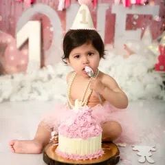 A fun and messy first birthday cake smash session. This little girl is enjoying her first taste of cake against a festive pink backdrop, a perfect way to celebrate a major milestone.
