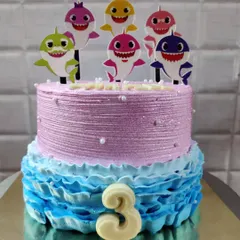 A fun two-tier Baby Shark cake for a third birthday. The bottom tier features blue buttercream waves, while the top is decorated with the whole Baby Shark family.