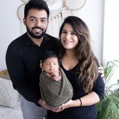 A classic family portrait with a newborn. The baby is swaddled and held securely by both parents, who are smiling warmly at the camera.
