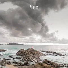 An epic, wide-angle shot of Harman and Komal standing on the rocks against a dramatic, cloudy sky in Phuket. This image showcases the power and beauty of a coastal landscape.