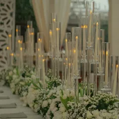 A long dining table set for an all-white wedding, featuring a runner of fresh white flowers and dozens of candles in tall glass hurricanes for a clean and elegant feel.