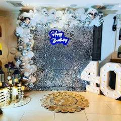 A glamorous 40th birthday photo booth with a silver sequin wall, a blue neon sign, and silver and white balloon decor.