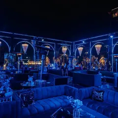A stunning Sangeet setup with dramatic blue lighting and crystal chandeliers. The plush velvet seating and moody ambiance create a luxurious and glamorous party setting.