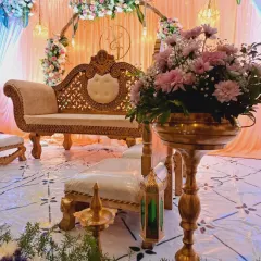 This engagement setup combines traditional and modern elements. The stage features a classic diwan-style sofa, traditional brass lamps (diya), and Moroccan-style lanterns, all set against a backdrop of soft peach and blue drapes with a floral arch.