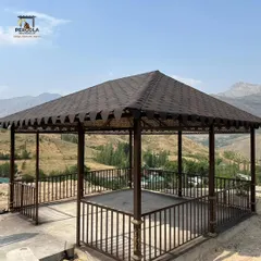 This magnificent shingle-roof gazebo is perched on a hillside, offering a breathtaking view. The intricate metalwork on the railings and eaves adds a touch of decorative elegance to the sturdy structure.