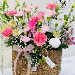A charming purse-shaped wicker basket filled with a delicate arrangement of pink carnations and white accent flowers. This unique design is a whimsical and stylish way to gift flowers.