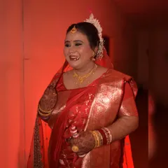 A candid photograph of a Bengali bride sharing a laugh. Dressed in a traditional red saree and holding a 'gach kouto', this image captures a moment of pure, unscripted joy.