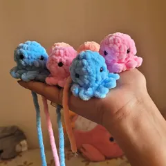 A group of my fluffy octopus plushies in the making, before their keyrings are attached. Held in my hand, you can see their size and the vibrant pink, blue, and peach colors I use.
