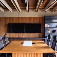 The Managing Director's cabin at Anthea Pharma features a discussion table set against a wood-paneled wall with integrated screens. The ceiling's cement board baffles conceal all lighting, maintaining a clean, mid-century modern aesthetic while wooden acoustic panels enhance sound quality.