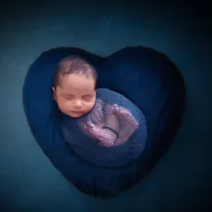 A beautifully swaddled newborn is curled up in the center of a plush, heart-shaped blue pillow, symbolizing a heart full of love.