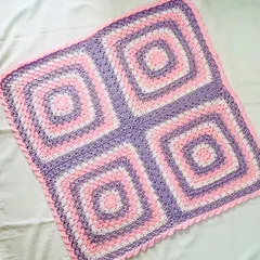A lovely granny square blanket in a classic four-square pattern, using soft pink, purple, and white yarn. The simple scalloped border adds a sweet, delicate finish.