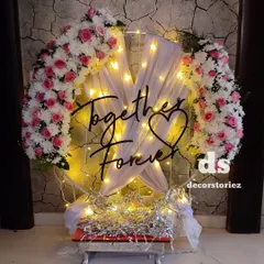 An elegant ring backdrop we created for a 25th anniversary. The design features a 'Together Forever' sign, draped white fabric, fairy lights, and lush arrangements of pink and white artificial flowers.