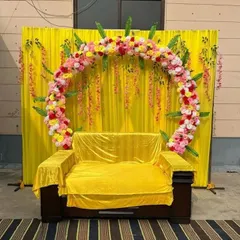 A simple yet beautiful Haldi seating area with a bright yellow fabric backdrop. A circular floral arch made of pink, yellow, and white flowers frames the covered sofa, making it a perfect spot for the ceremony's rituals and photos.