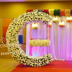 A stunning cradle ceremony setup featuring a crescent-shaped arch made of white flowers. The hanging cradle, also adorned with flowers, creates a beautiful focal point, while colorful lanterns add a festive touch.