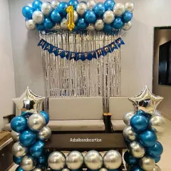 A clean and stylish first birthday decoration in blue and silver. The setup includes a balloon garland over a silver curtain backdrop, flanked by two balloon pillars topped with star-shaped foils.