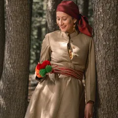 A beautiful solo portrait of the woman in her traditional Pahari dress, leaning against a pine tree. The details of her attire, like the colorful waist accessory, are highlighted.