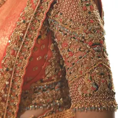 An extreme close up of an embroidered sleeve on an orange silk blouse. You can see the texture of the Zari thread, the shimmer of the beads, and the precision of each stitch.