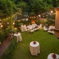 An intimate garden soirée setup, viewed from above. The combination of elegant white furniture, lush green lawn, and warm string lights creates a magical and inviting atmosphere for a small, sophisticated gathering.