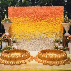 A beautiful floral wall with an ombre effect of orange, yellow, and white flowers. In the foreground, two large golden lotus-shaped urlis are ready for the Haldi ceremony, perfect for an outdoor garden setup.