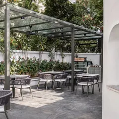The outdoor seating area at Kana Cafe, inspired by the breezy, nature-integrated cafes of Bali. The space features a glass canopy, minimal furniture, and a small arched opening that acts as a pickup point for delivery services, blending aesthetics with practicality.